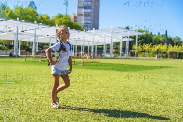 Young girl walking across a sunny green park field, carrying a small soccer ball and backpack,