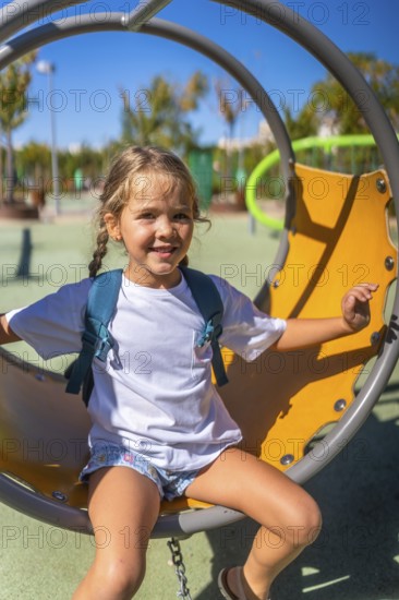 Happy little girl with a backpack smiles broadly while sitting on a swing in a vibrant outdoor