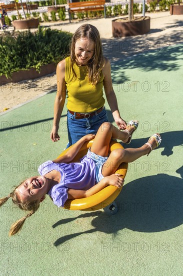 Mother standing, smiling, and gently pushing her happy daughter on a yellow spinning toy, both