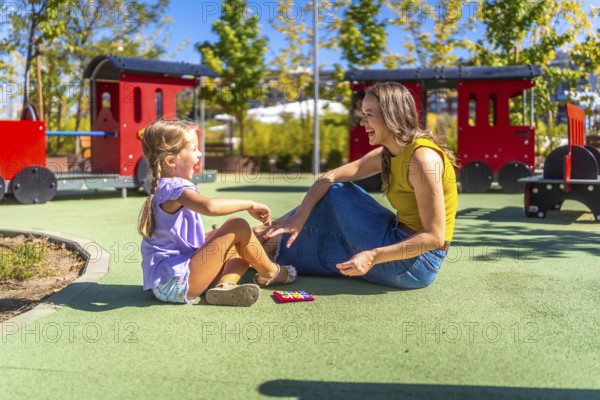 Mother and daughter sitting on soft flooring at a public playground, laughing happily during a
