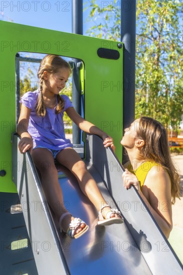 Mother encouraging her smiling daughter on a sunny park slide, sharing a playful bonding moment of