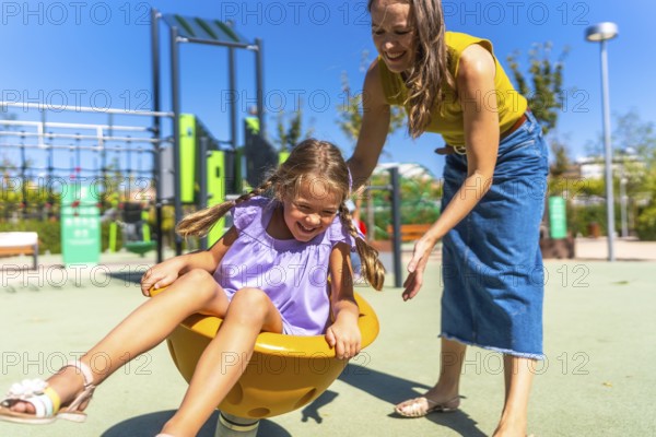 Mother and daughter laughing on a colorful spring rider at a sunny outdoor playground, sharing a