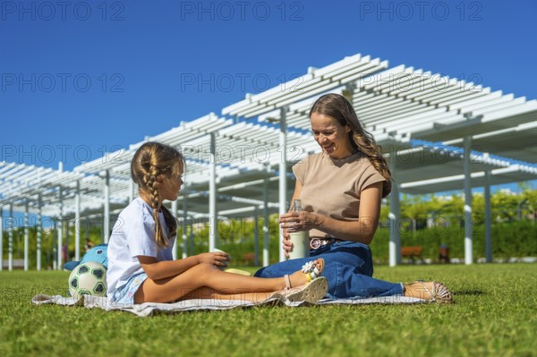 Mother and daughter sitting on a picnic blanket in a sunny urban park, smiling and bonding over a