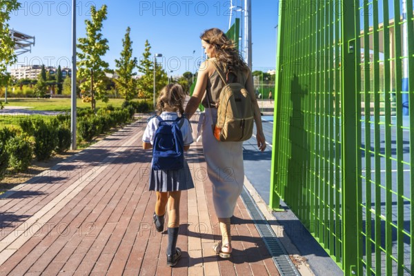 Mother and daughter walk hand in hand on a sunny sidewalk, child in school uniform and backpack