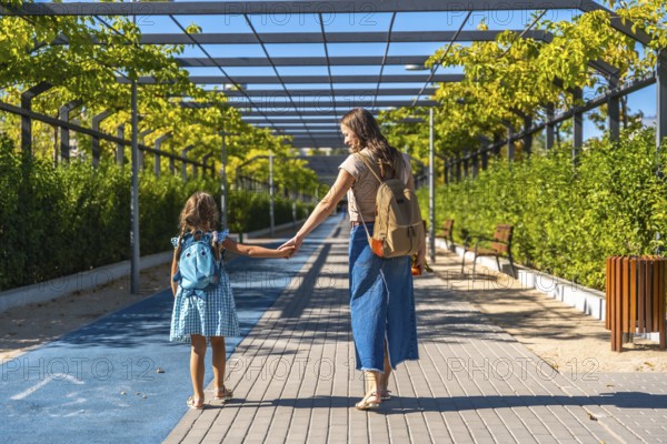 Mother and daughter walking along an urban path, holding hands and carrying backpacks on a sunny