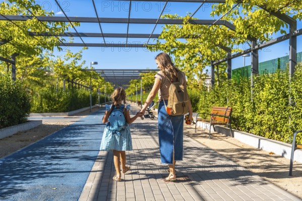 Mother and daughter, wearing backpacks, walking along a sunny pathway lined with green trees and