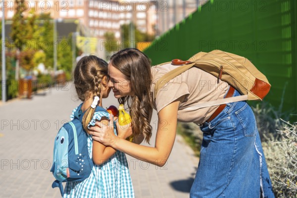 Loving mother bending down to embrace her small daughter with a backpack on the first day of