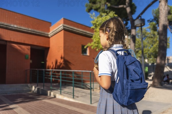 Young schoolgirl wearing a uniform and carrying a blue backpack, walking towards the entrance of an