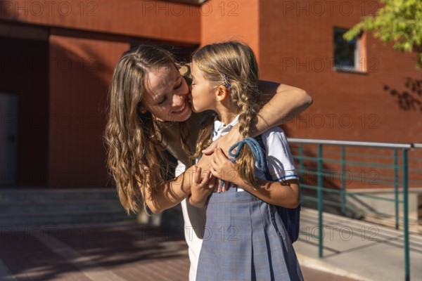 Mother embraces and kisses her young daughter in school uniform at the school entrance on the first