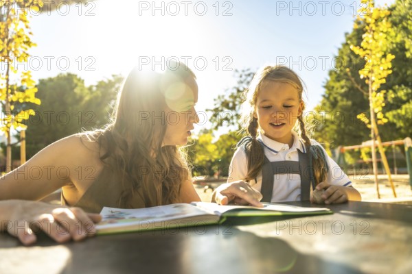 Mother and daughter bonding over reading a book together, sitting at a table outdoors in a bright