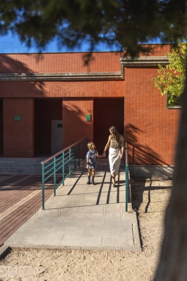 Mother and daughter holding hands and walking together, approaching a school building on a sunny