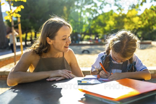 Mother watching her daughter drawing and doing homework at an outdoor table at school, enjoying a