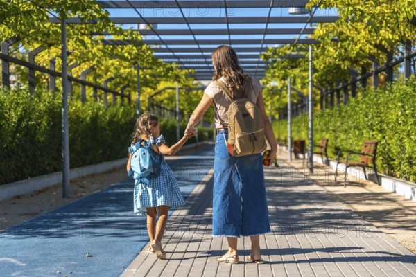 Mother and daughter holding hands and walking together along a paved path, carrying backpacks and