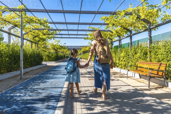 Mother and daughter walk hand in hand along an urban park path with backpacks, heading to school on