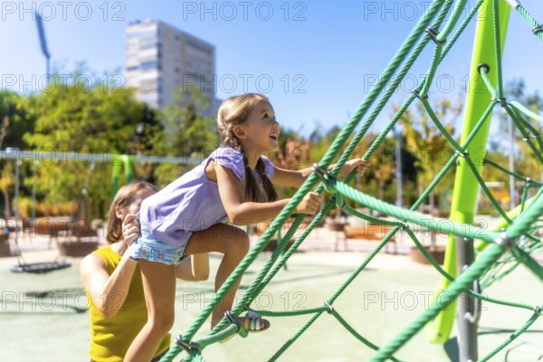 Mother assisting her happy daughter climbing a green rope jungle gym on a sunny day, experiencing