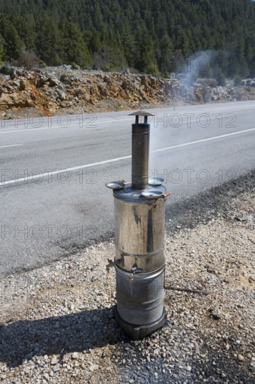 Smoking metal stove with roadside chimney against wooded backdrop, traditional samovar, kettle for