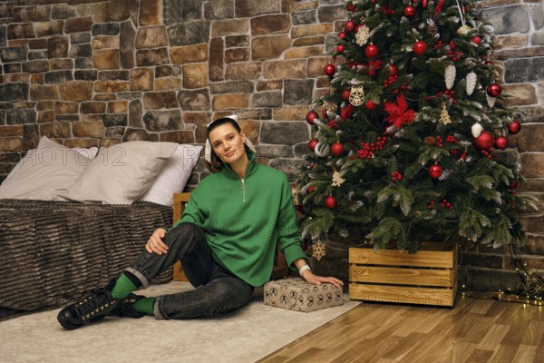 A cheerful woman sits on the floor beside a beautifully decorated Christmas tree in a cozy indoor