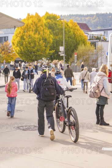 A man pushes his bike across a sidewalk while people and autumn trees form the backdrop,