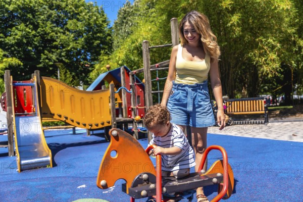 Young mother assisting her son while playing on a rocking horse in a colorful playground on a sunny