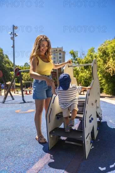 Happy mother assisting her toddler son while he's climbing stairs on a playground structure,