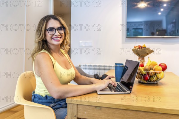 Smiling woman using laptop at kitchen table, working remotely with fruit basket and headphones