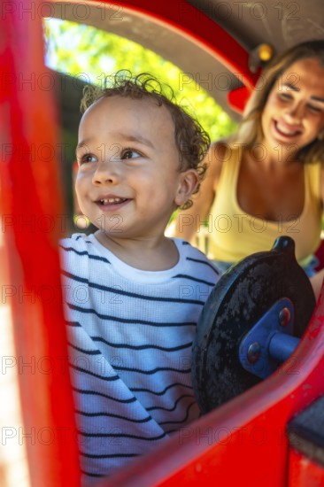 Smiling toddler playing joyfully on a playground slide while mother watches nearby, creating