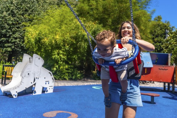 Young mother having fun with her baby son, pushing him on a swing in a colorful playground during a