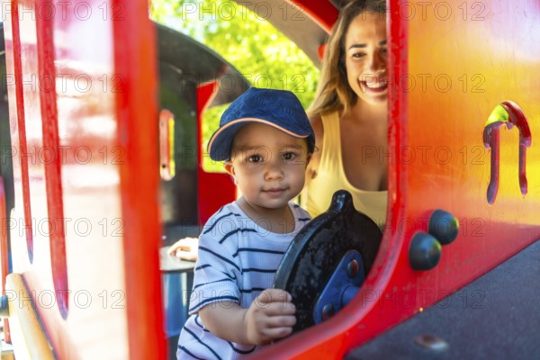 Happy toddler having fun driving a toy train at the playground while his mother watches him with a