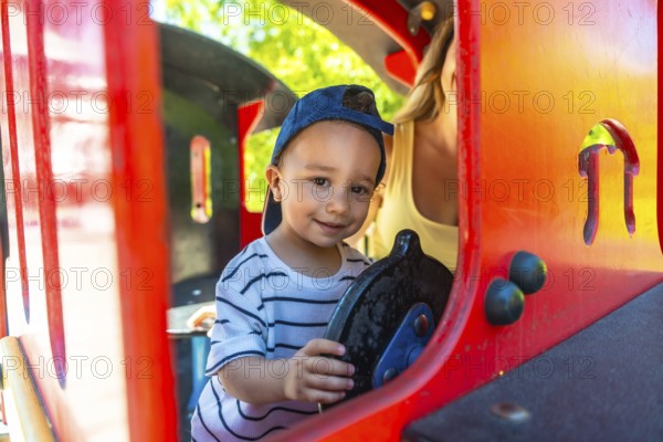 Little boy having fun driving a toy train at the playground, with his mother supervising in the