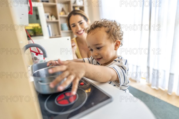 Happy toddler boy having fun playing with a toy kitchen while his mother watches over him in a