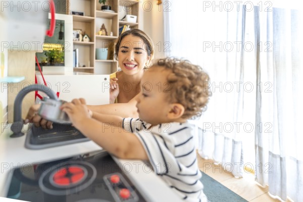 Smiling mother looking at her little son having fun playing with a toy kitchen in a modern, bright