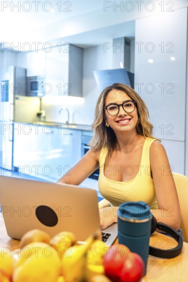 Young woman working on laptop in modern kitchen with fresh fruits, promoting healthy work life