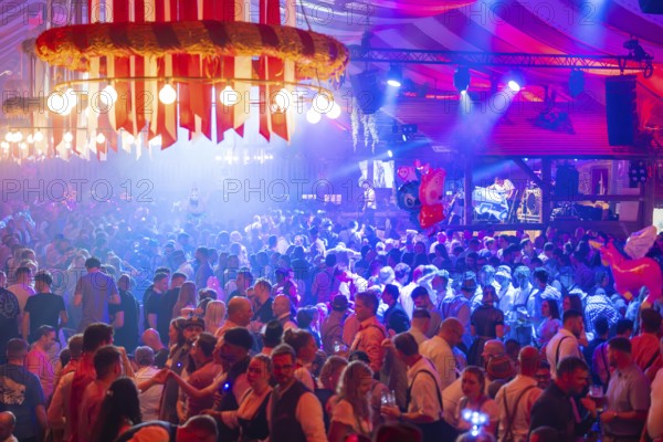 A lively party in a festive beer tent with stage and illuminated atmosphere at night, Volksfest