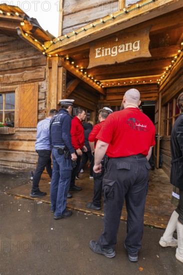 People stand in front of a wooden entrance, some wearing red shirts. A security-conscious