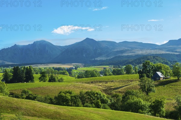 Lush green fields with a solitary house, set against majestic mountains and a bright sky, Massif of