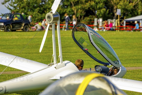 A motorised glider parked at the edge of the airfield with the canopy open during an air show at