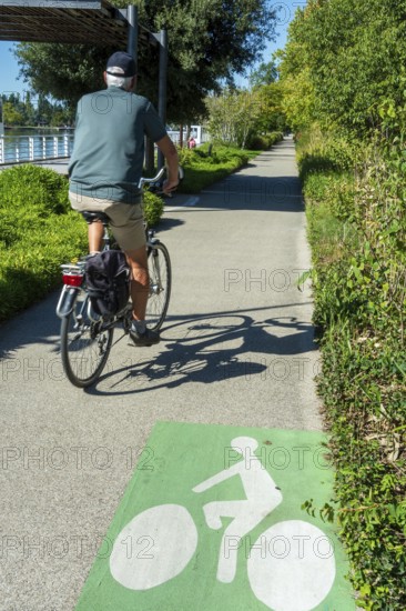 A person rides a bicycle on a smooth pathway lined with trees and shrubs. The sun shines bright,