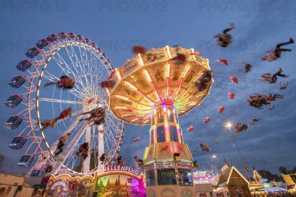 An illuminated chain carousel and Ferris wheel at night at a funfair, people swinging on chains,