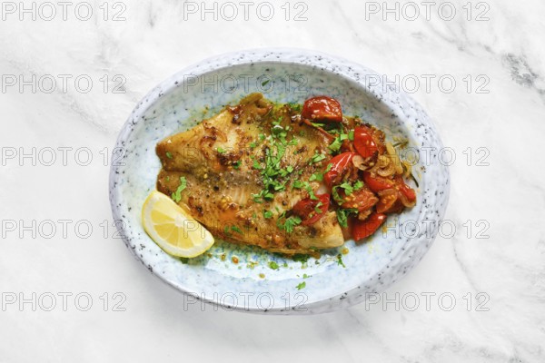 A perfectly cooked white fish fillet is topped with sautéed tomatoes and fresh herbs. A lemon wedge