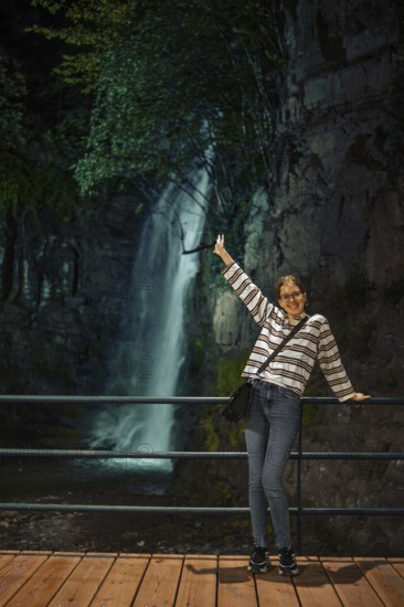 A cheerful young woman stands on a wooden walkway behind a railing, posing joyfully with one arm