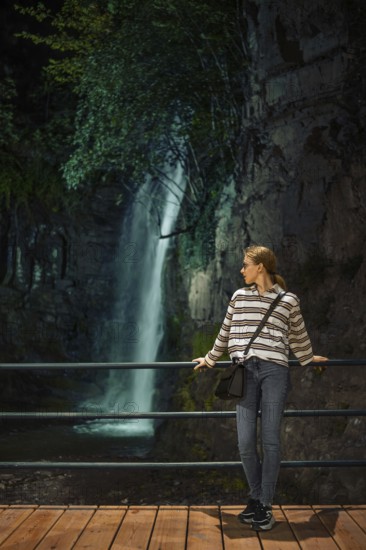 A young woman enjoys a quiet moment by a waterfall in a lush, natural setting during the evening