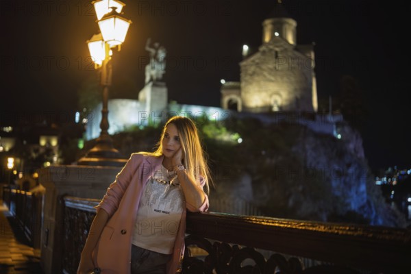A woman leans against a railing at night, dressed casually with a light jacket. Behind her, a