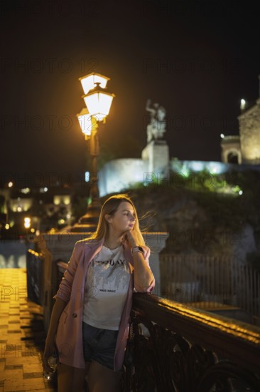 A woman stands by a decorative railing of the bridge, admiring the night scenery. Dim lights