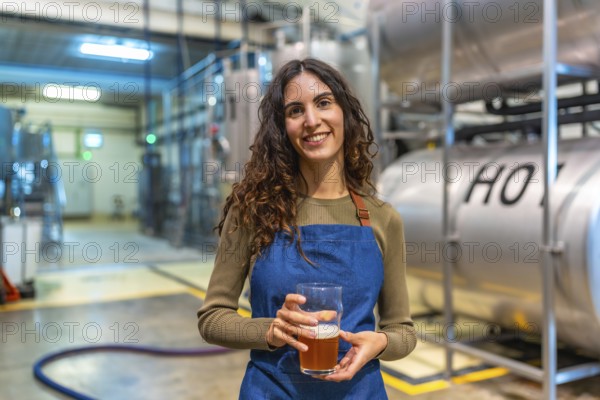 Woman brewer standing in modern brewery, smiling and holding a glass of craft beer for quality