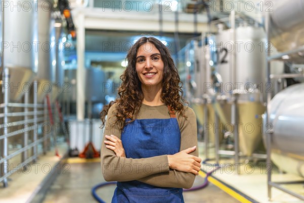 Woman brewer stands confidently with arms crossed, smiling at camera in a modern microbrewery with