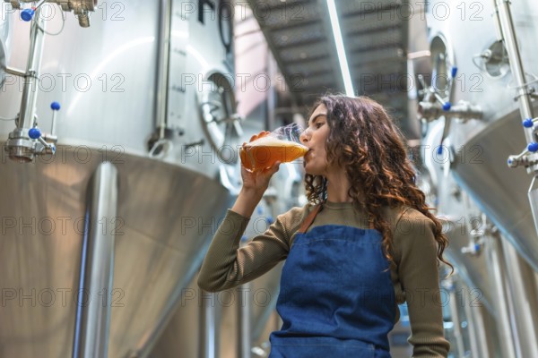 Woman wearing an apron tasting a glass of freshly brewed beer for quality control, standing in a