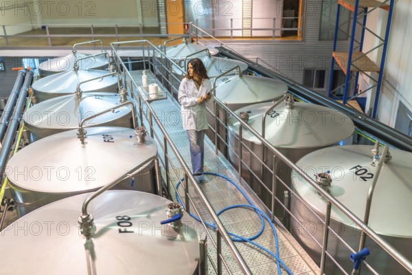 Woman scientist in a lab coat walking on a platform among large industrial fermentation tanks,