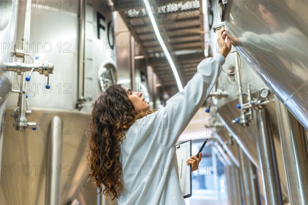 Young woman in a lab coat checking control panel on a stainless steel fermentation tank, performing