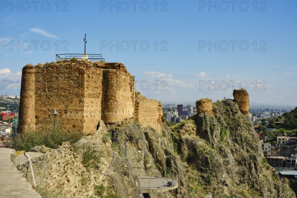 A weathered stone fort stands on a rocky hill, crowned with a cross. Below, a vibrant city