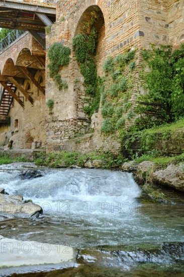 Clear water streaming over smooth stones next to an ancient stone wall adorned with lush plants.